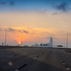 The orange sun rises through a haze of clouds behind the OKC skyline with four lanes of highway in the foreground.